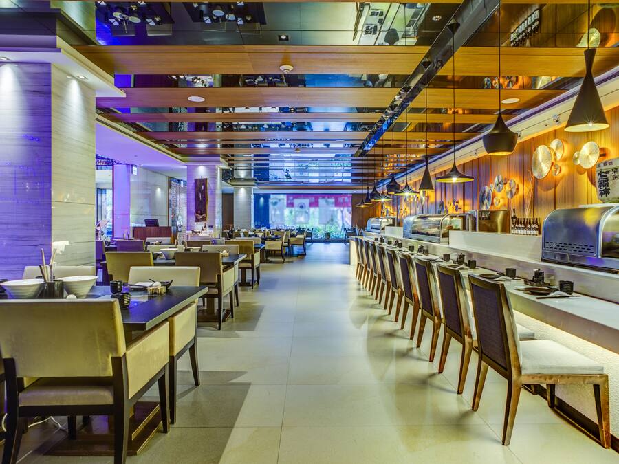 A modern restaurant with multiple dining tables and chairs, a long counter with a display case, and hanging pendant lights.