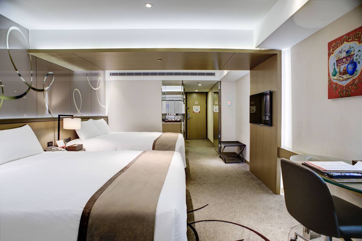 A hotel room with two beds, a desk, a chair, a TV, and a bathroom visible in the background.