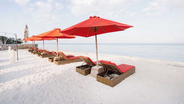 Ein Strand mit Liegestühlen und roten Sonnenschirmen, ein klarer Himmel und ein ruhiges Meer.