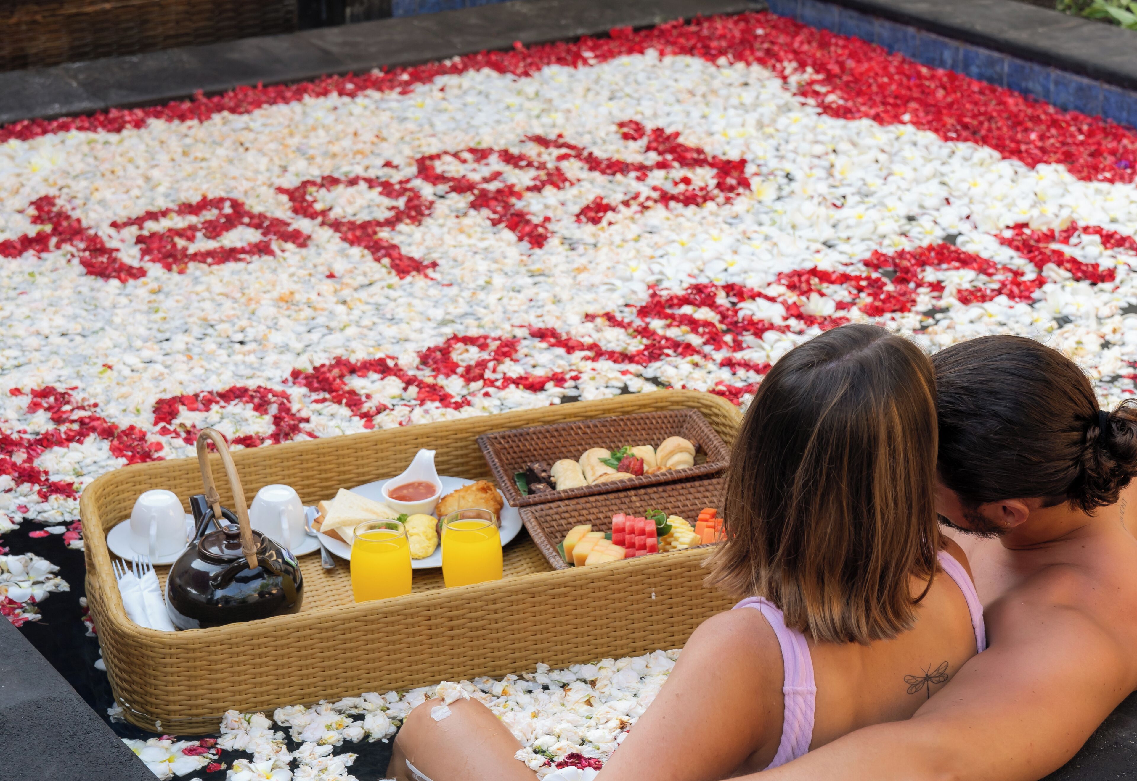 Un couple est assis sur un lit recouvert de pétales de fleurs, avec un plateau de petit-déjeuner devant eux.