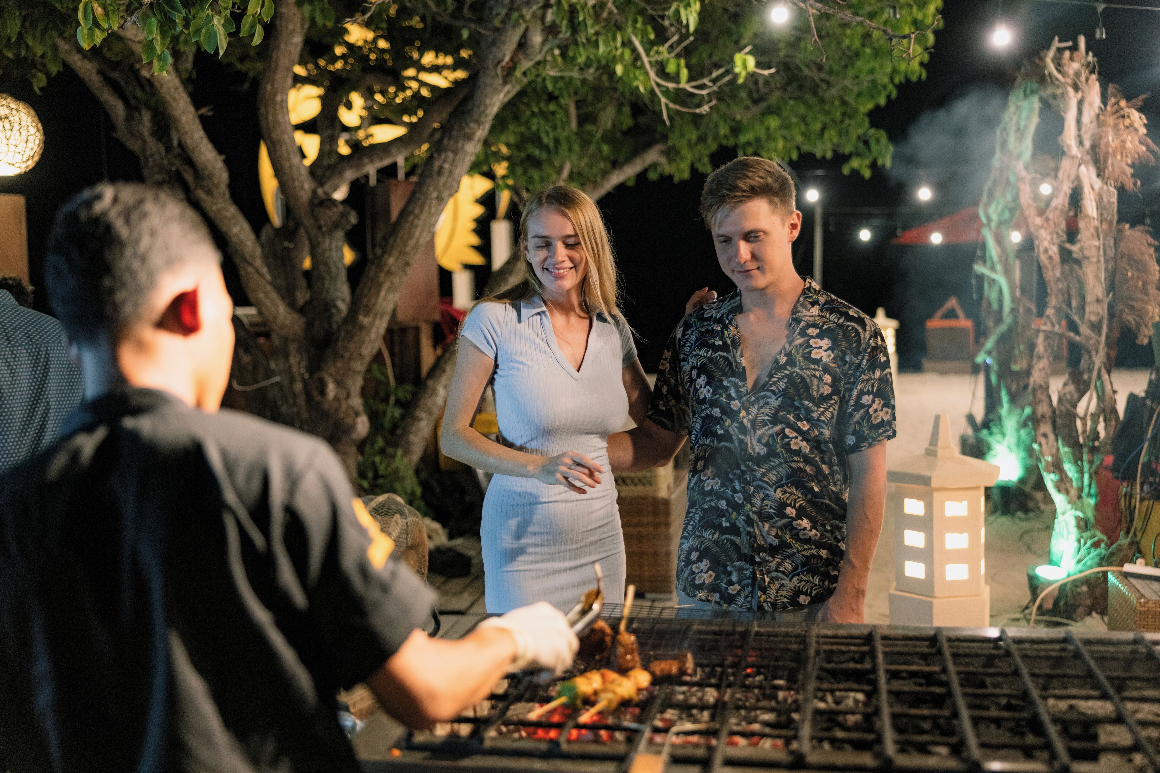 Eine Gruppe von Menschen versammelt sich um einen Grill, im Hintergrund Bäume und dekorative Lichter.