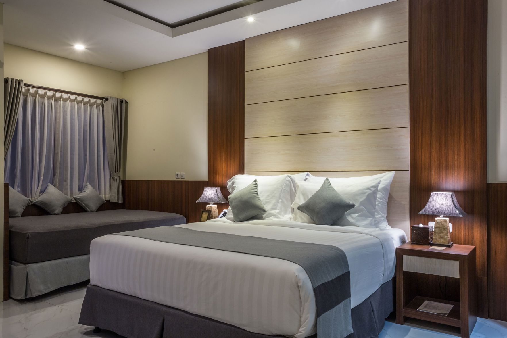 A hotel room with a large bed, two bedside tables, a nightstand, a window with curtains, and a wooden headboard.