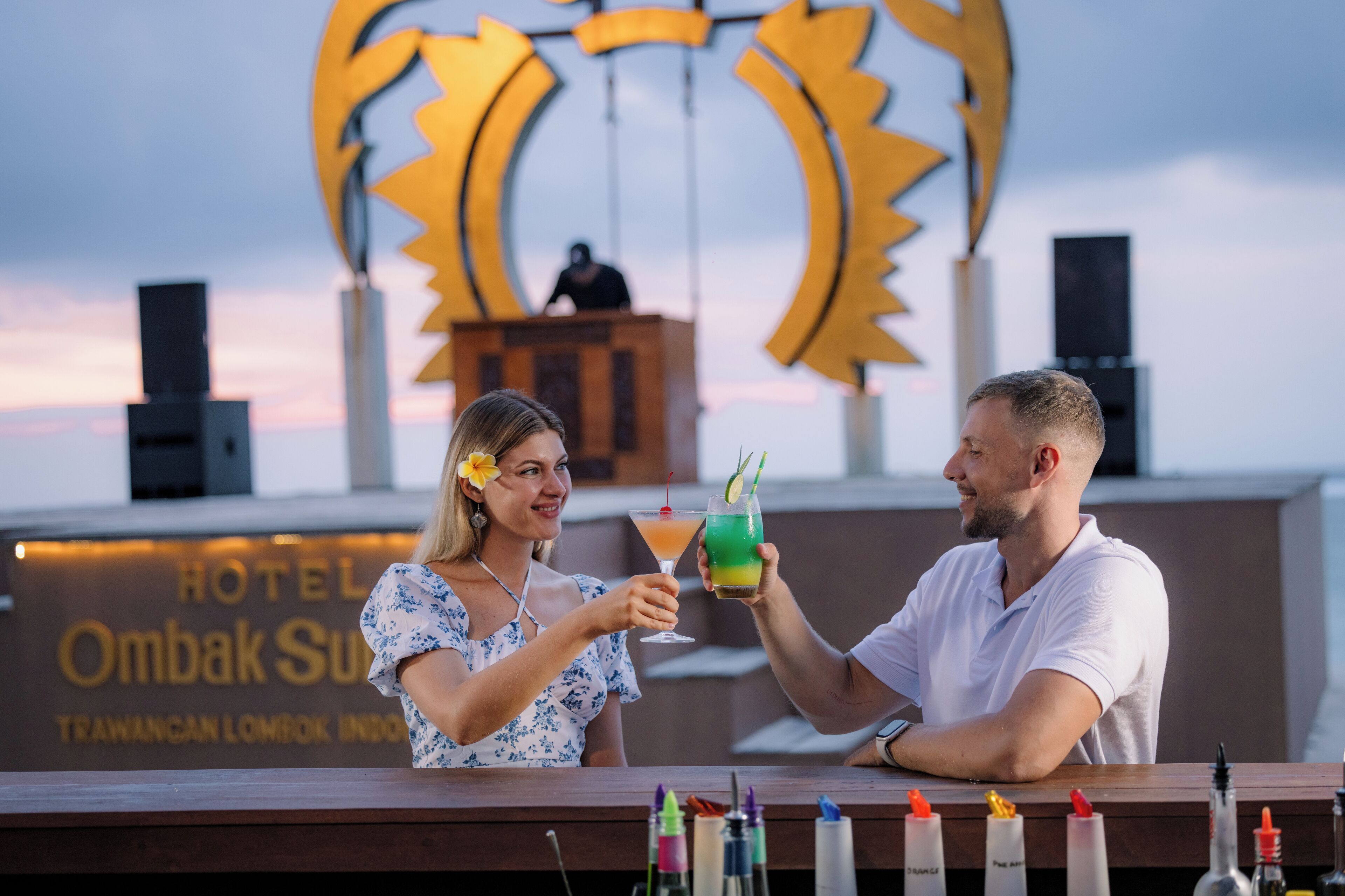 Two people toasting with drinks at a bar with a sign reading "HOTEL OMBAK SUN" in the background.