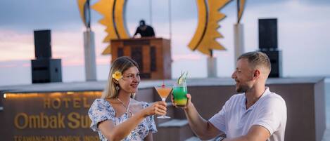 Two people toasting with drinks at a bar with a sign reading "HOTEL OMBAK SUN" in the background.