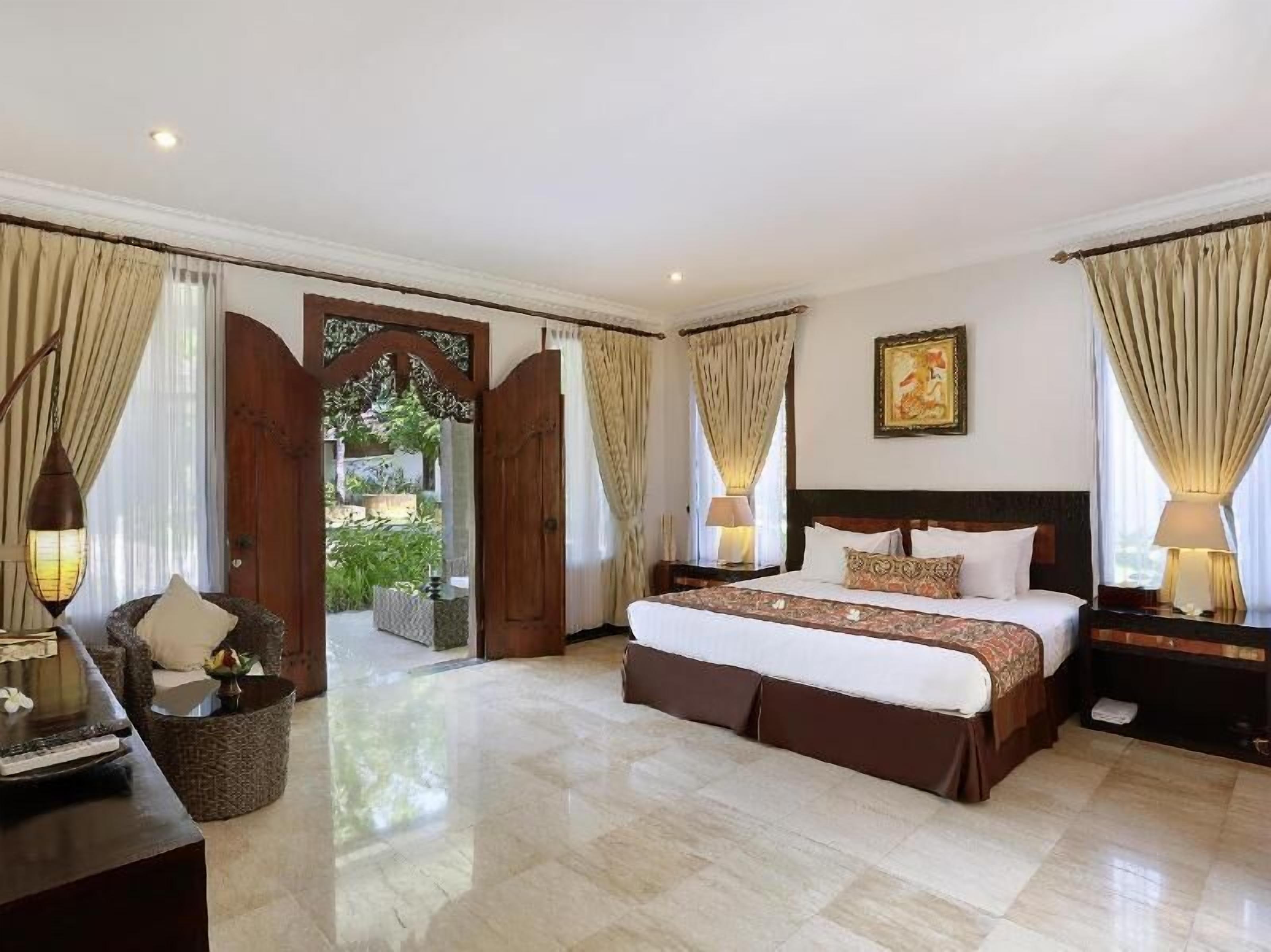 A spacious bedroom with a large bed, a nightstand, a chair, and a view of a garden through an open door.