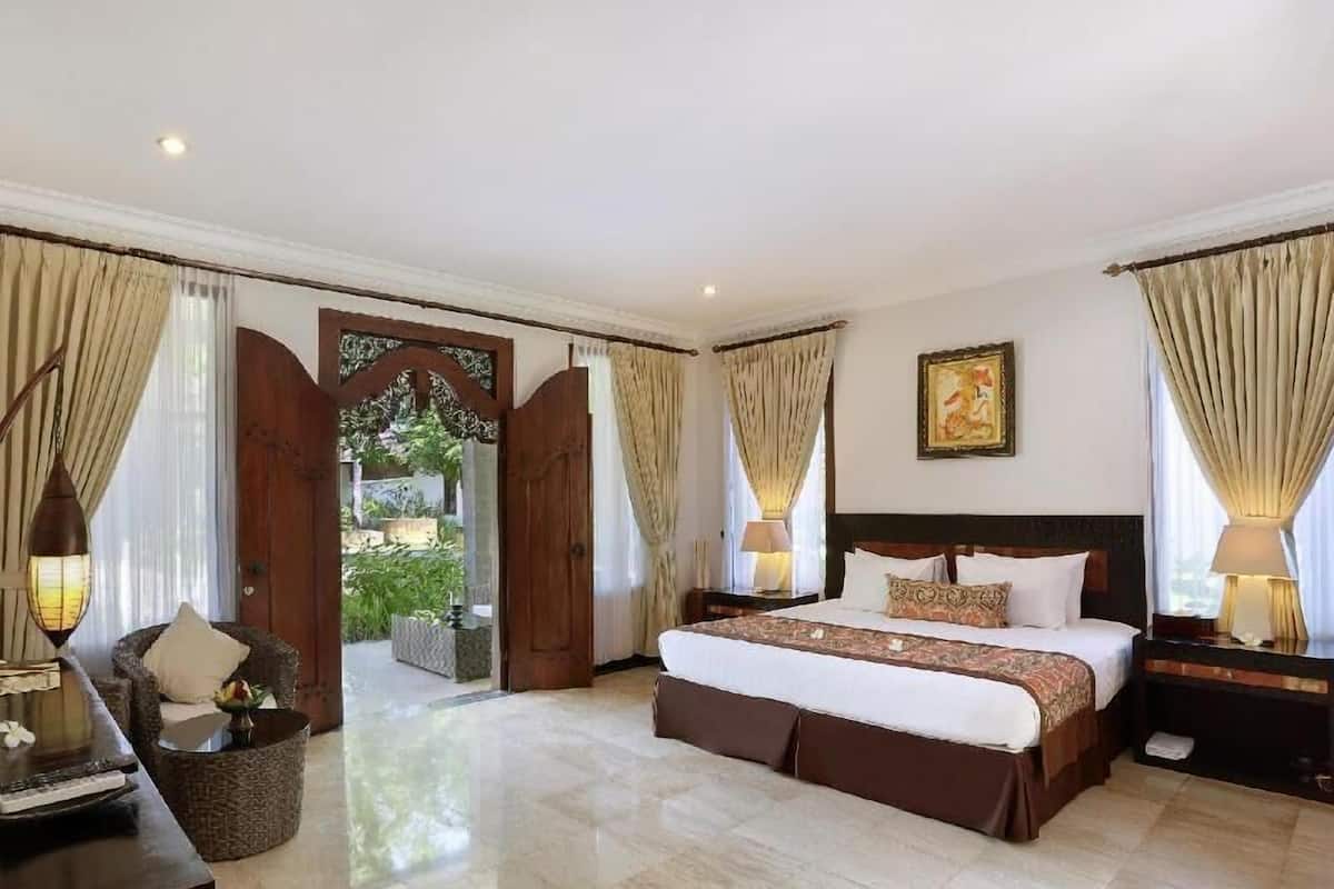 A spacious bedroom with a large bed, a nightstand, a chair, and a view of a garden through an open door.