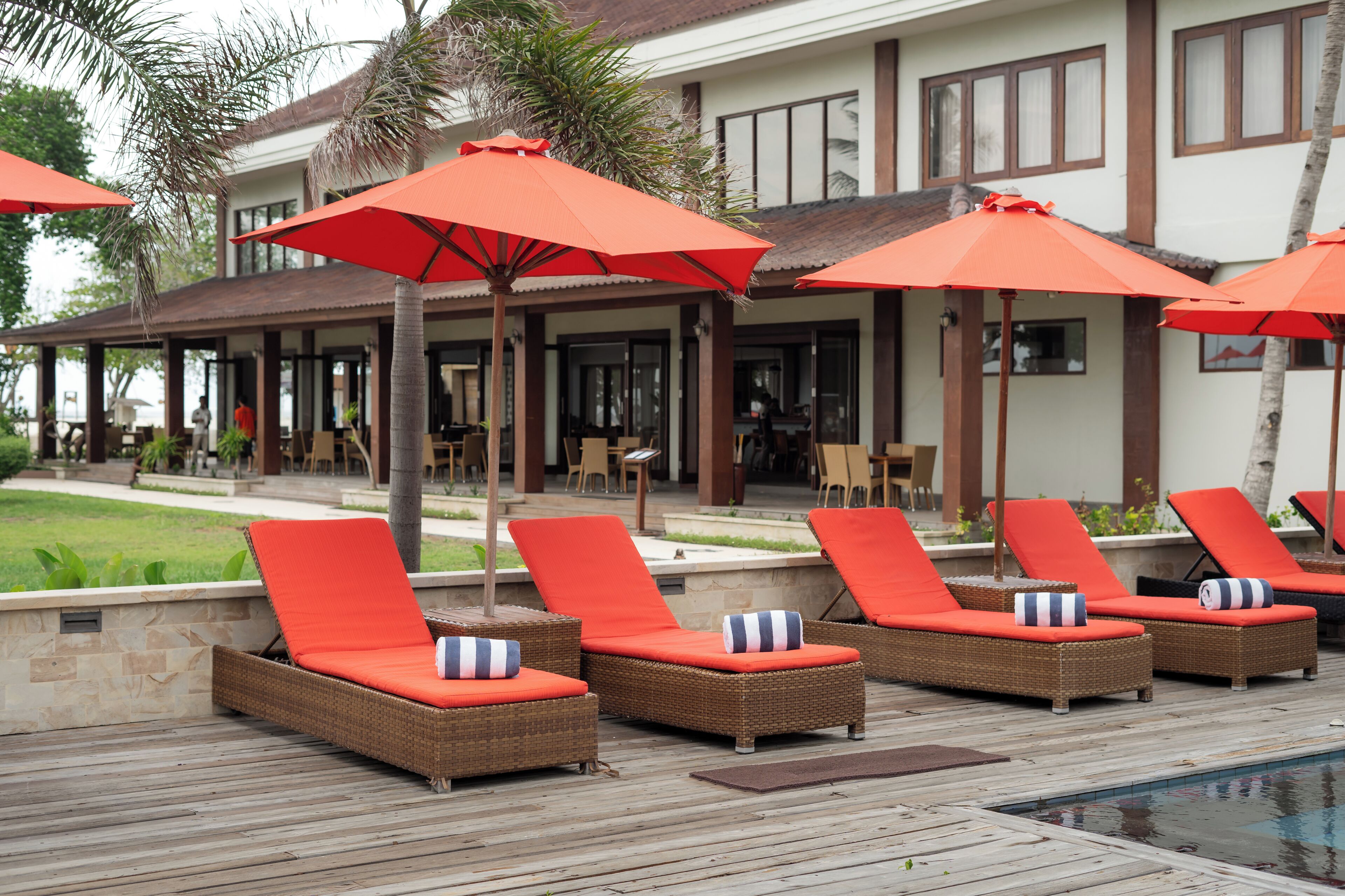 Ein Poolbereich mit Korlounge-Stühlen und roten Sonnenschirmen, einem Pool und einem Gebäude mit großen Fenstern im Hintergrund.