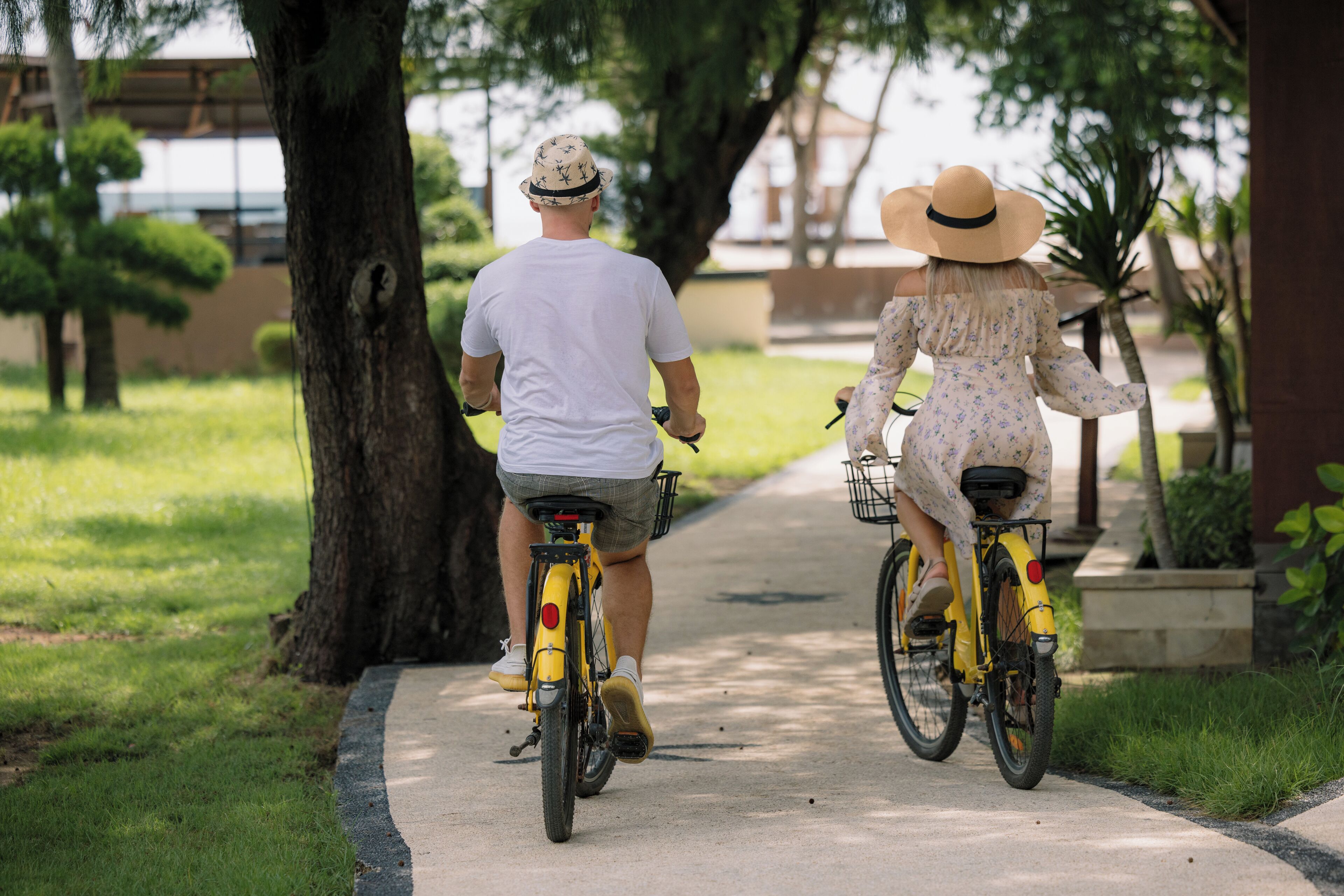 Dos personas andando en bicicleta por un camino pavimentado en un parque con árboles y césped.