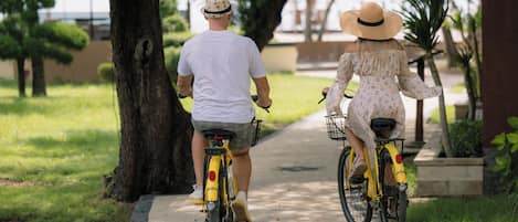 Dos personas andando en bicicleta por un camino pavimentado en un parque con árboles y césped.