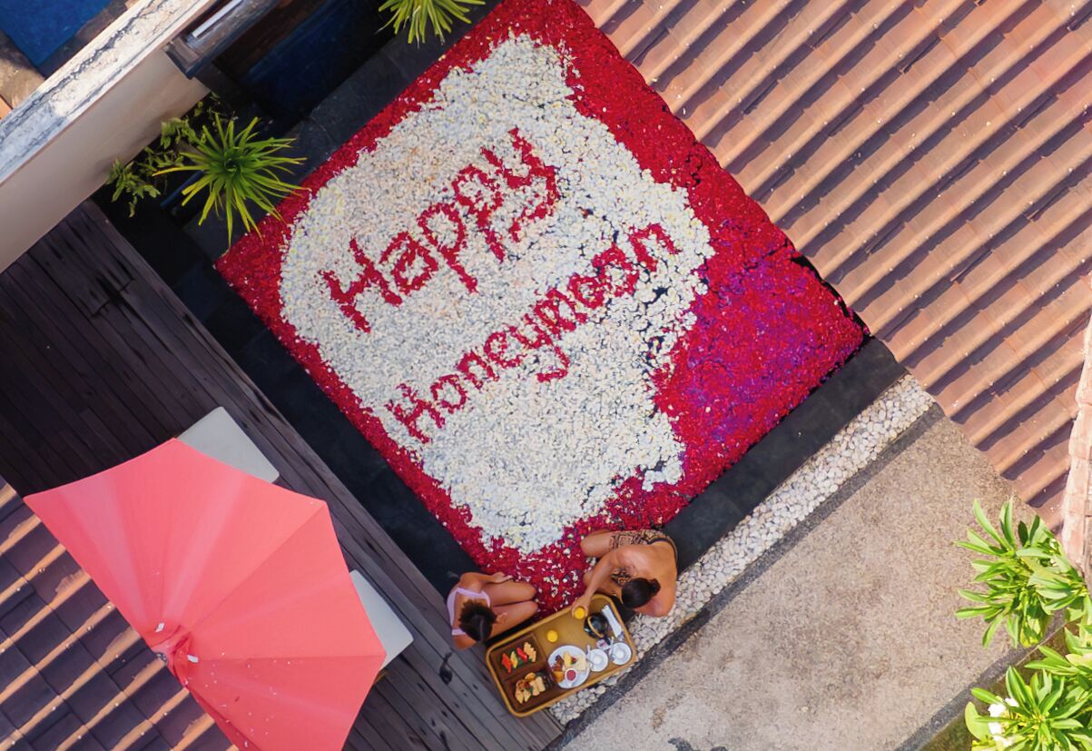 A welcome mat with "Happy Honeymoon" written on it, placed on a wooden deck with a red umbrella and a small table with a drink.