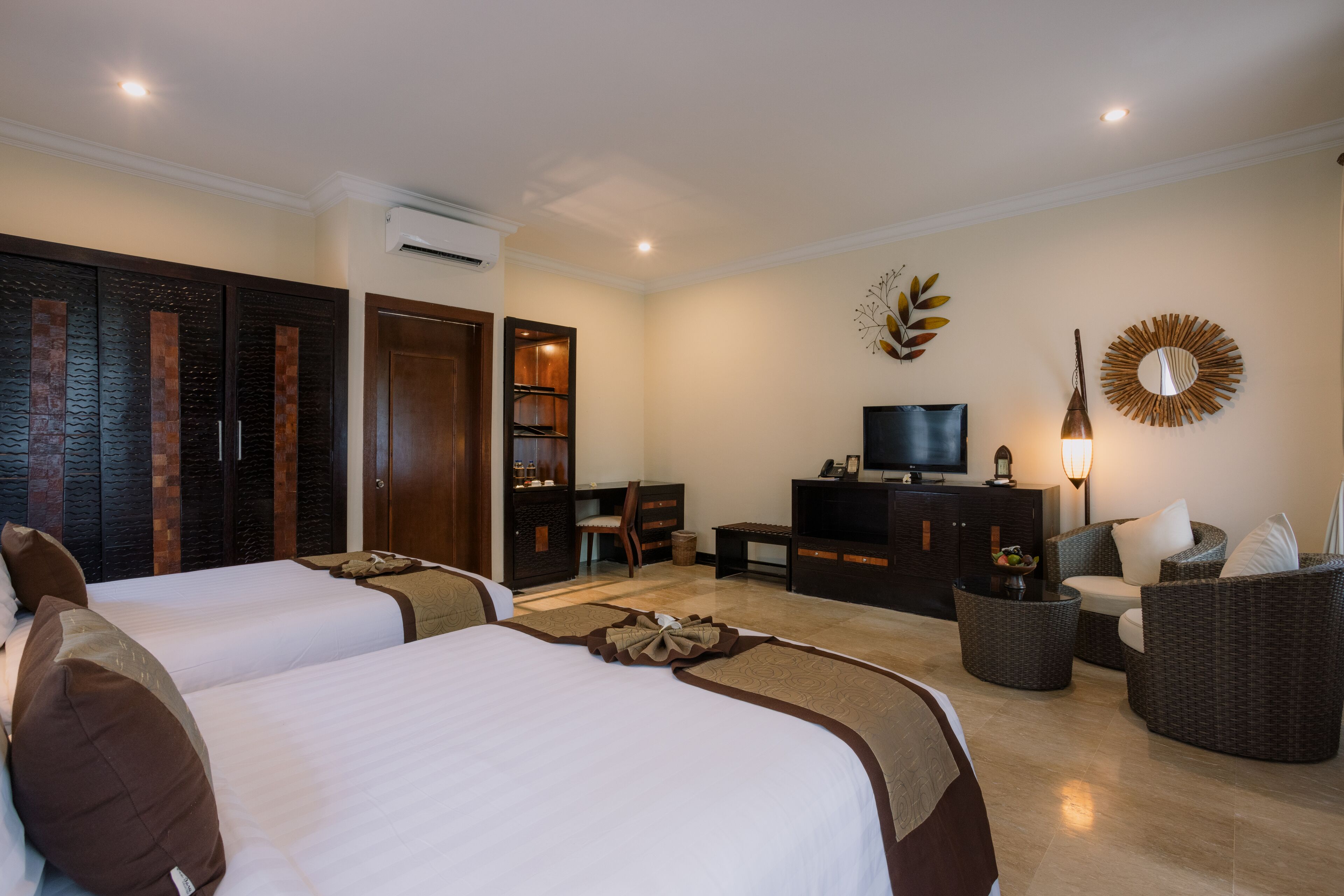 A hotel room with two beds, a desk, a television, and a seating area with a lamp.