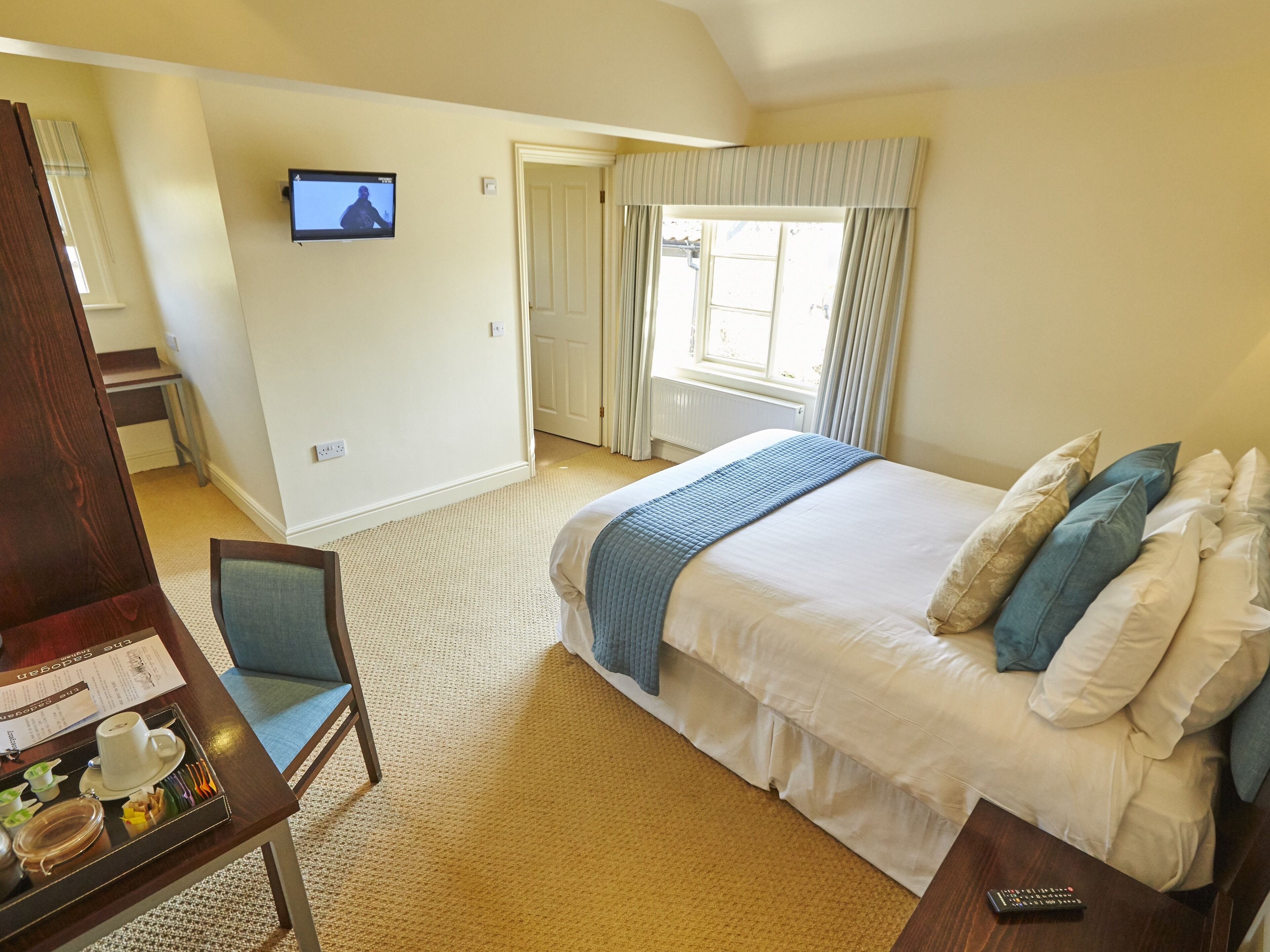 A hotel room with a bed, a desk with a tray of breakfast items, a TV on the wall, and a window with curtains.