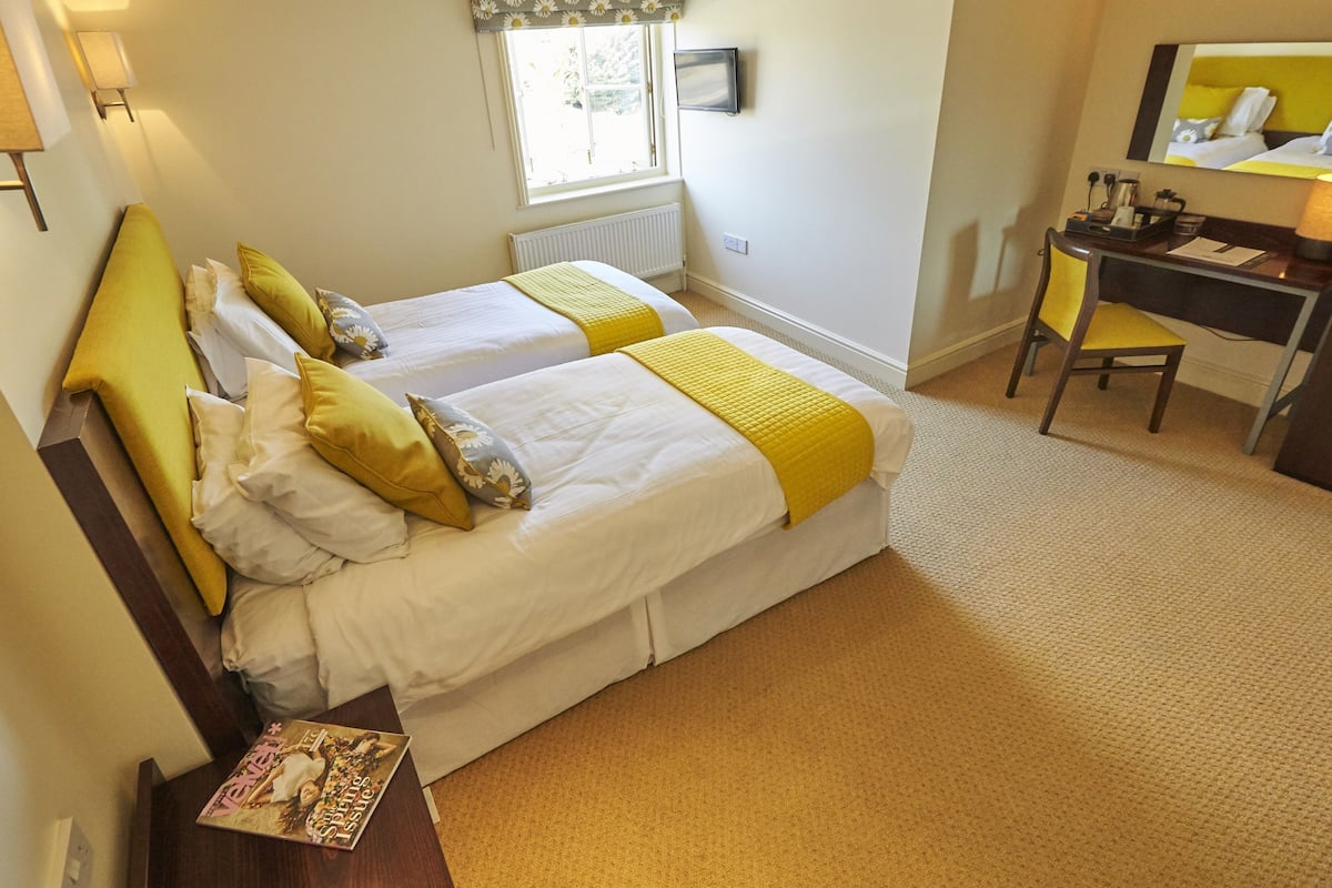 A hotel room with two beds, a desk, a chair, a radiator, a window with a flower-patterned valance, and a television on the wall.