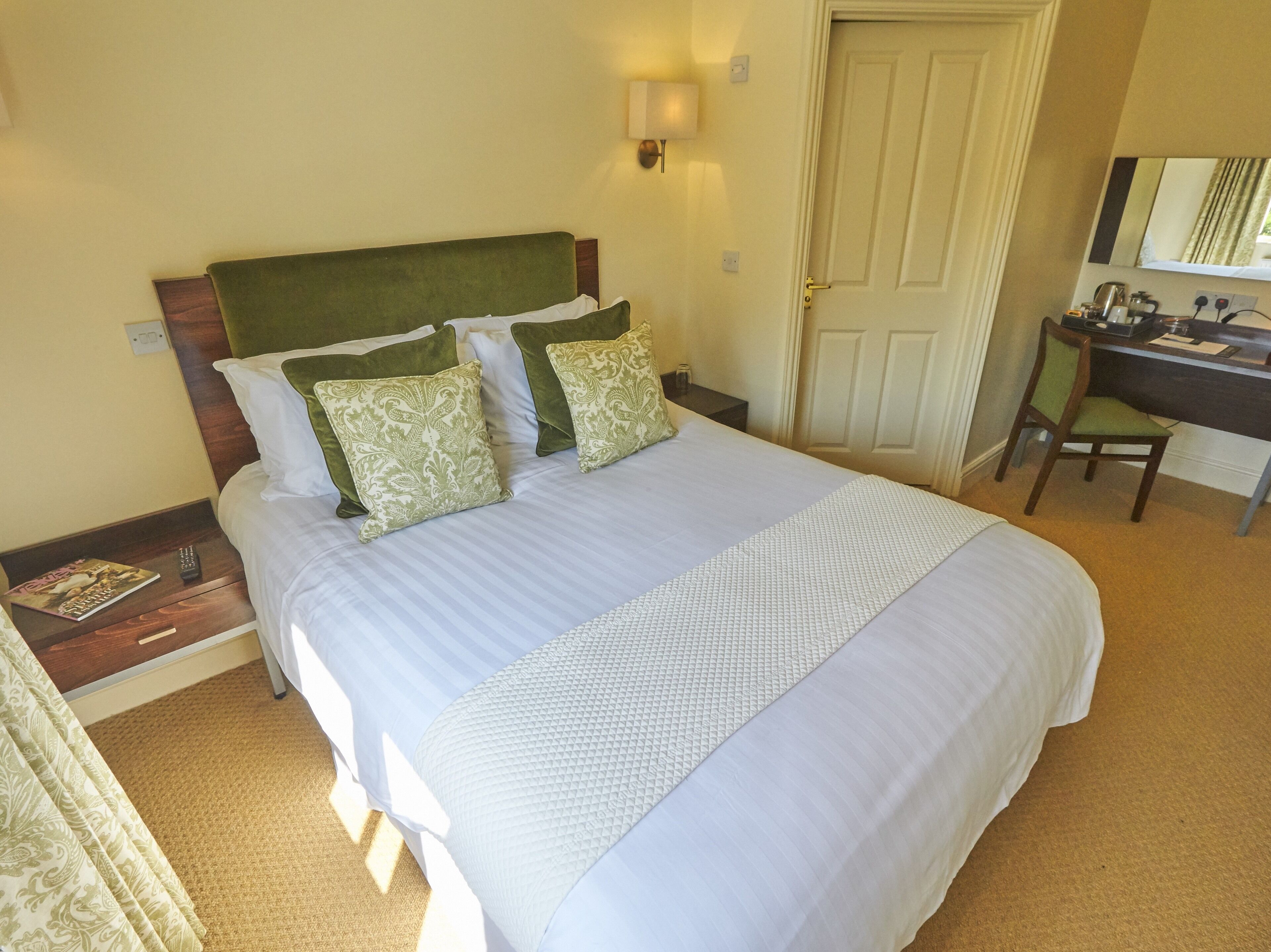 A neatly made bed with a green headboard, two patterned pillows, and a white bedspread. A desk with a chair and a lamp is visible in the background.