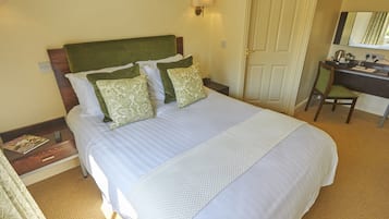 A neatly made bed with a green headboard, two patterned pillows, and a white bedspread. A desk with a chair and a lamp is visible in the background.