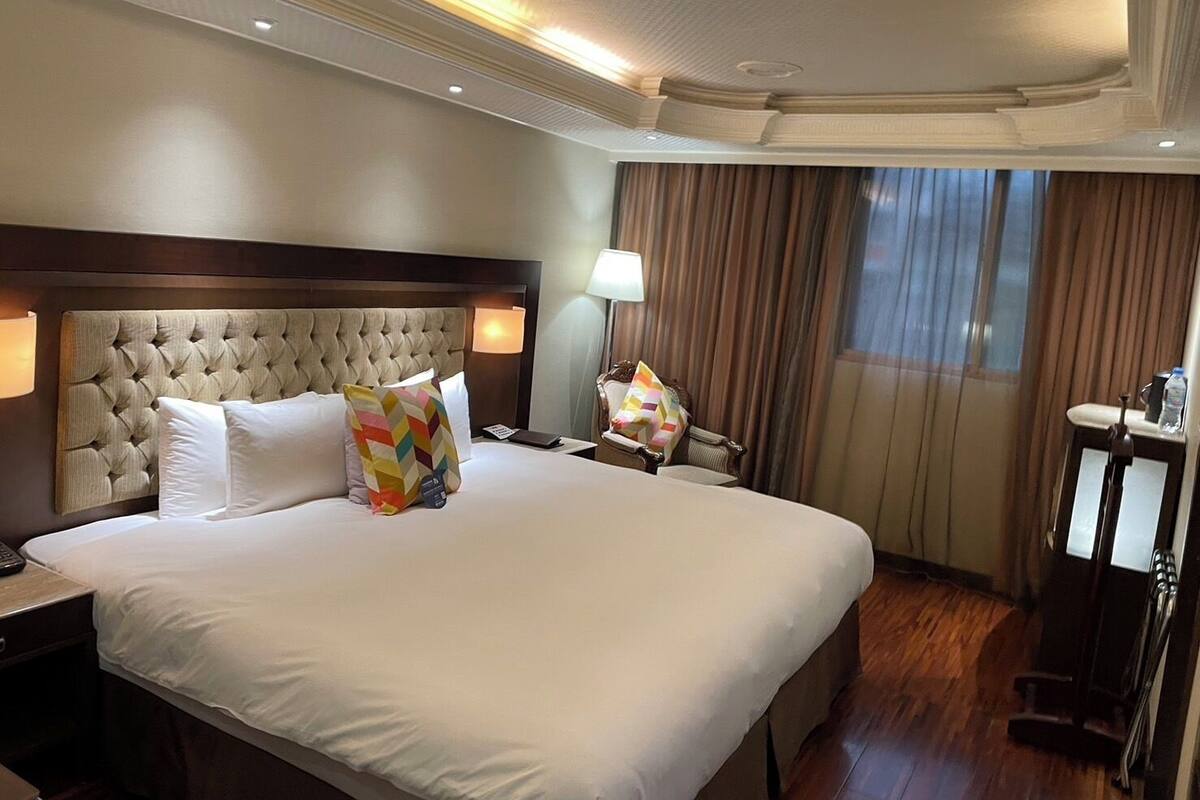 A hotel room with a large bed, a headboard, a nightstand, a lamp, a chair, a desk, and a window with curtains.