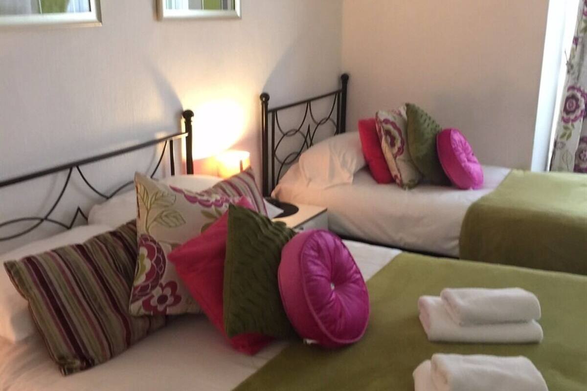 A hotel room with two beds, each with a green bedspread and multiple pillows. There are white towels on the beds.