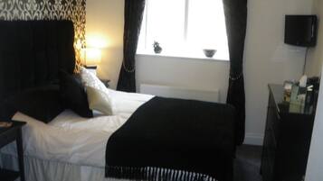 A bedroom with a bed, a nightstand, a lamp, a window with curtains, a television, and a radiator.