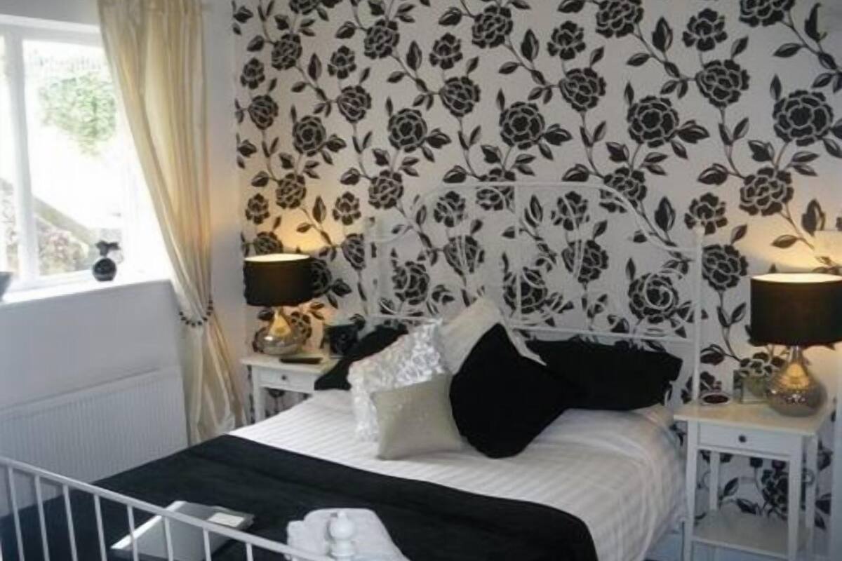 A bedroom with a bed, two bedside tables, a window with curtains, and a floral wallpaper pattern.