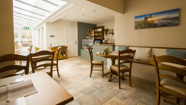 A dining area with a high ceiling, skylight, and a view of the outdoors. There is a kitchen area with a refrigerator and shelves.