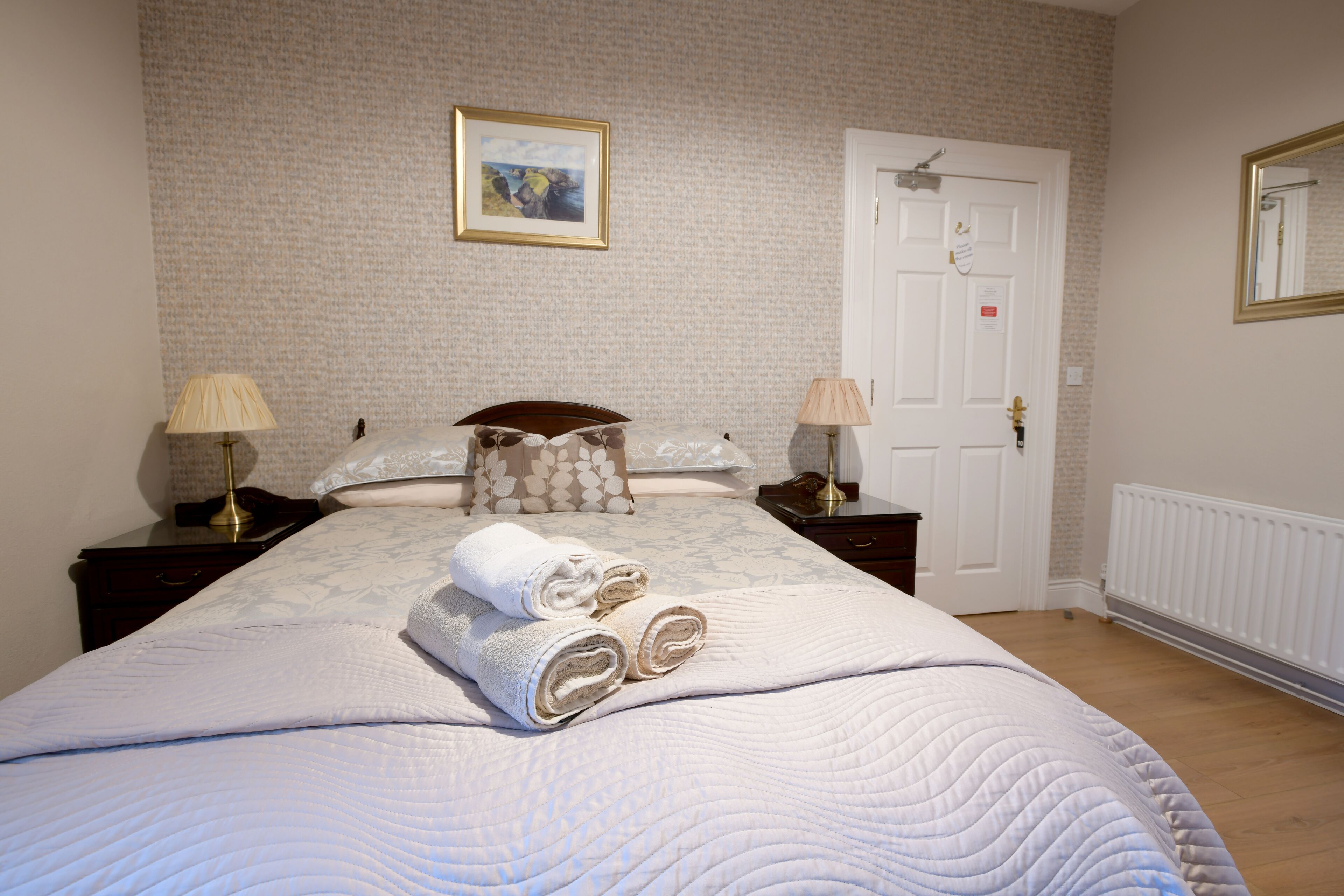 A neatly made bed with rolled towels, two bedside tables with lamps, a framed picture on the wall, and a white door.