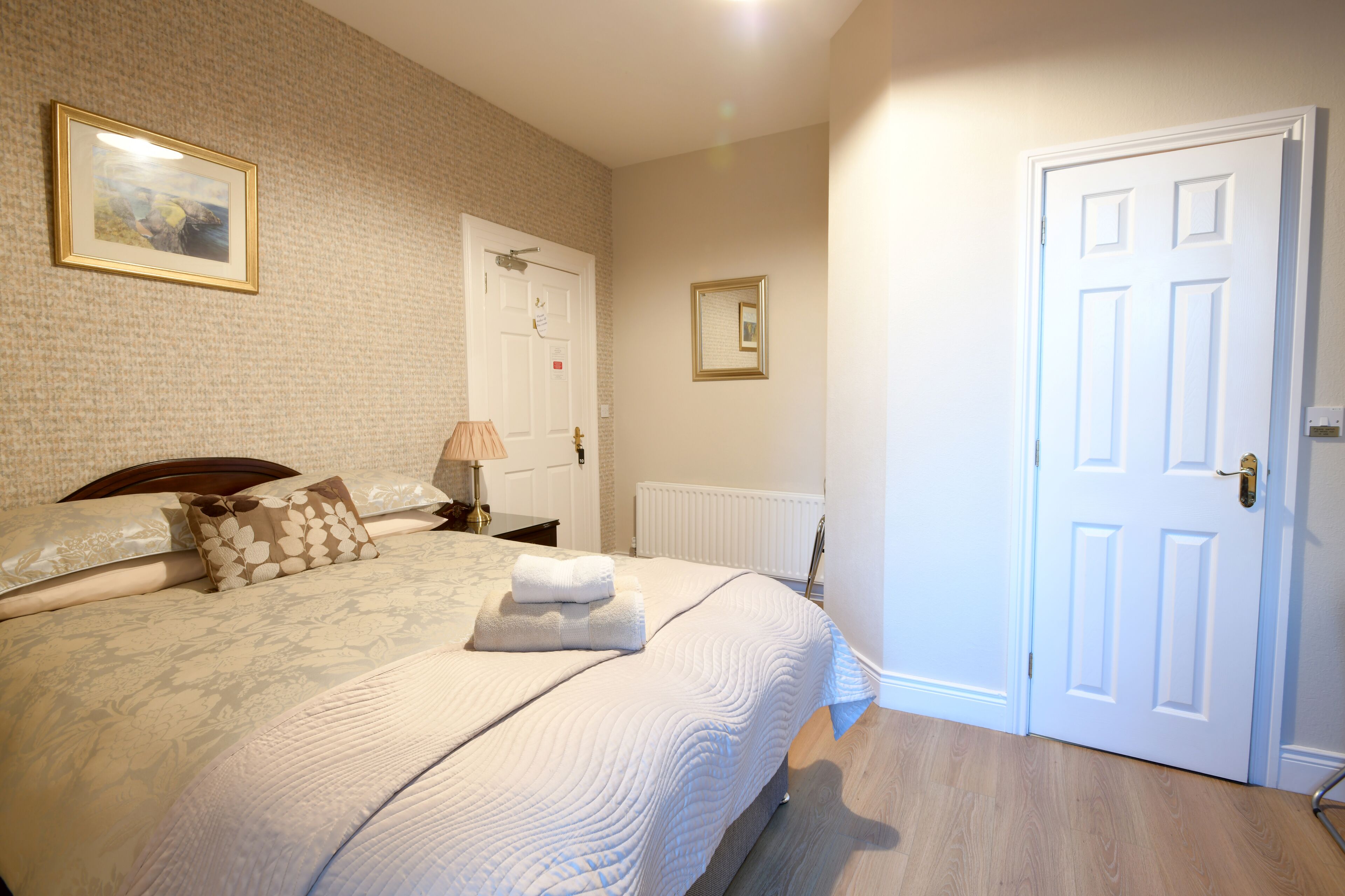 Een netjes opgemaakt bed met een witte deken en kussens, een nachtkastje met een lamp, en een ingelijst beeld aan de muur.