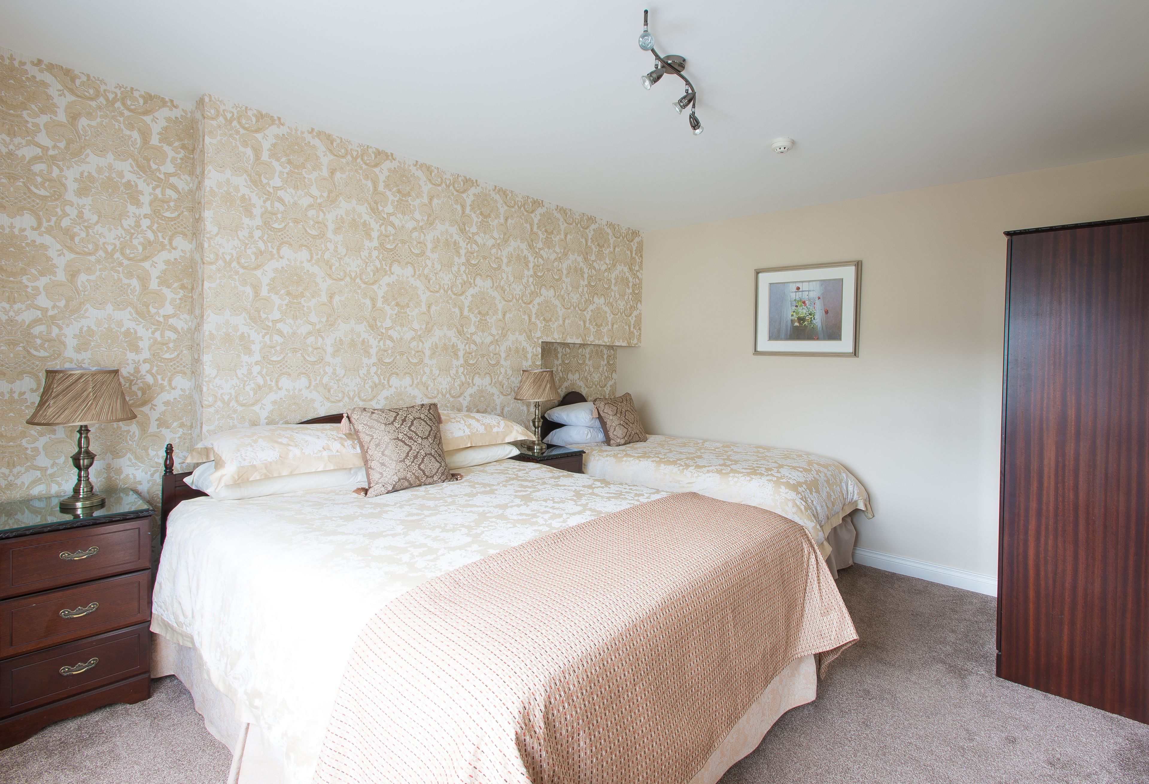 A bedroom with two beds, a dresser, a nightstand, a lamp, a framed picture on the wall, and a wooden wardrobe.