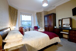 Quarto de hotel com uma cama grande, escrivaninha, cadeira, guarda-roupa e janela com cortinas.