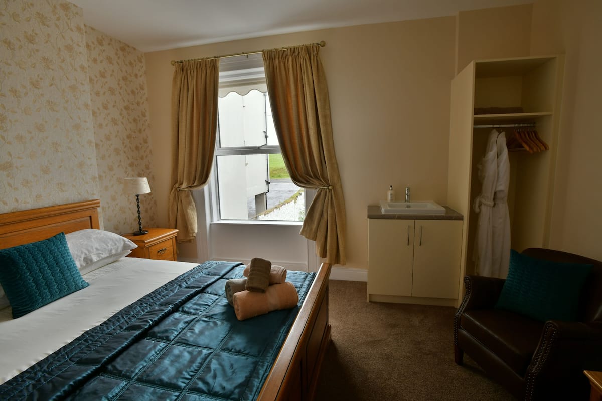 Habitación de hotel con cama, mesita de noche, ventana con cortinas, armario y baño con lavamanos.