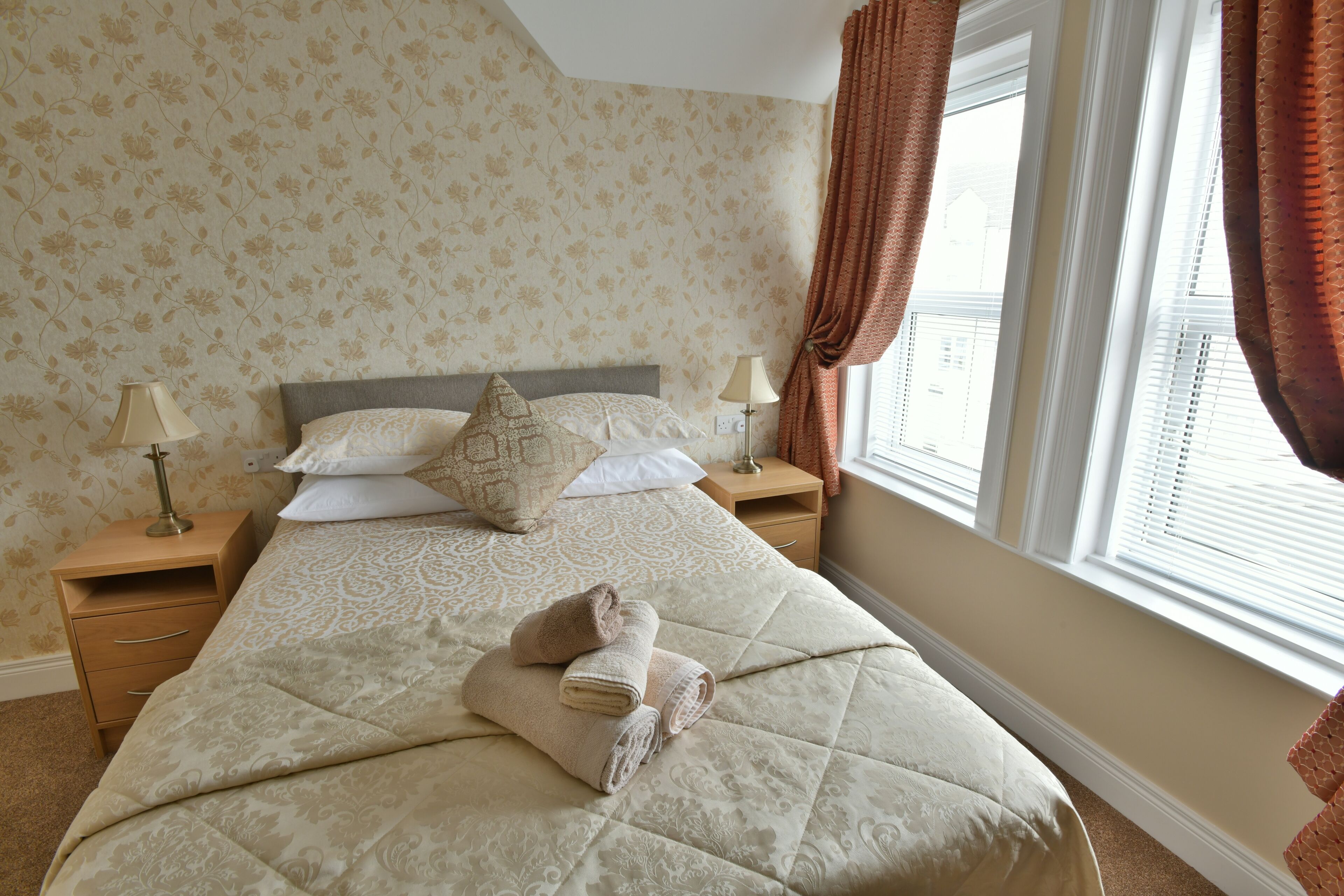 Una cama bien hecha con colcha, dos mesitas de noche con lámparas, una ventana con cortinas y un estampado floral en el papel tapiz.