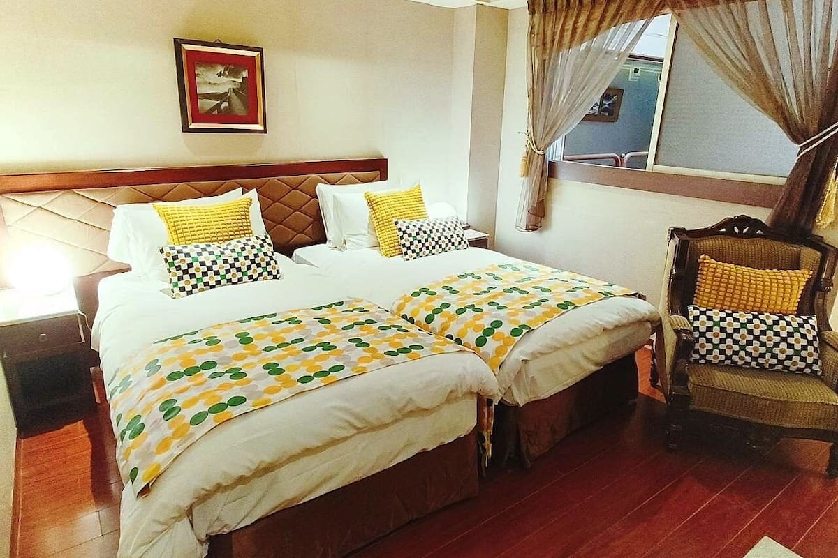 A hotel room with a double bed, a wooden headboard, a chair with a cushion, a framed picture on the wall, and a window with curtains.