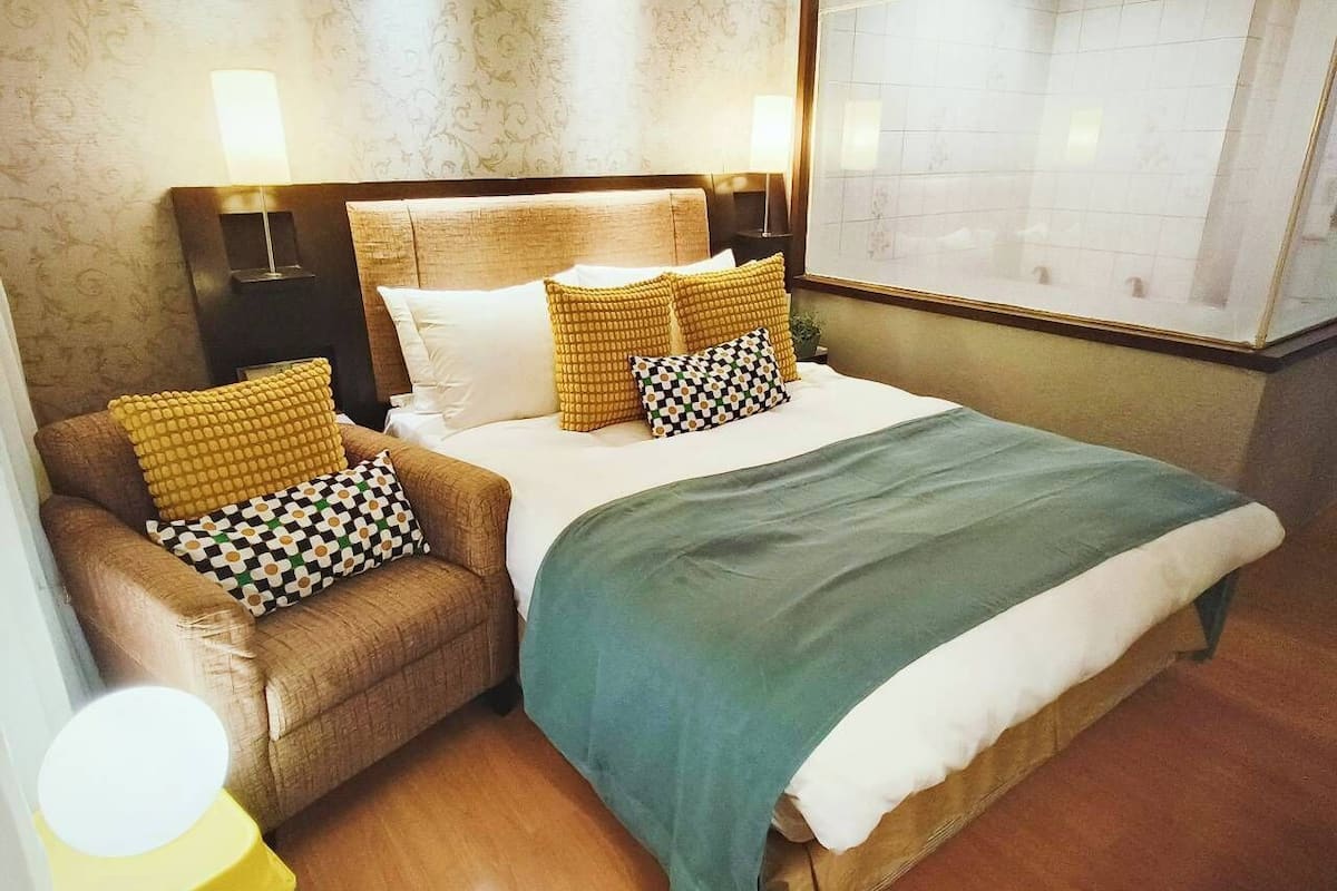 A hotel room with a large bed, two bedside tables with lamps, a small sofa with patterned cushions, and a trash bin.