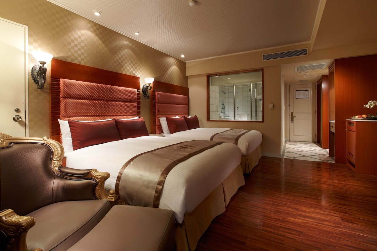 A hotel room with two large beds, a wooden floor, a bathroom with a glass shower, and a small desk with a plant.