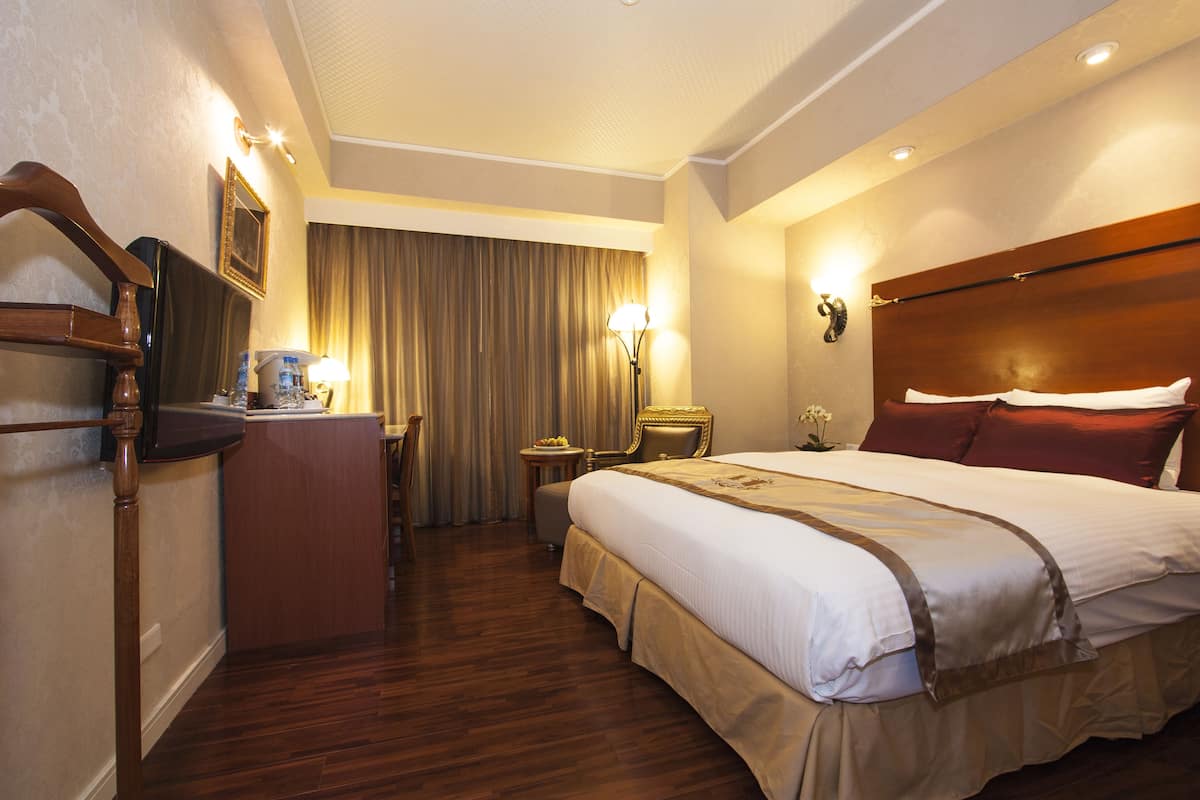 A hotel room with a large bed, a desk with a television, a chair, a lamp, and a curtain.