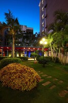Un patio de hotel bien iluminado al anochecer con piscina, palmeras y un gran parterre de flores.