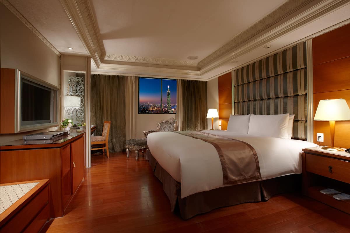 A hotel room with a large bed, a desk with a TV, a chair, and a view of a cityscape at night.