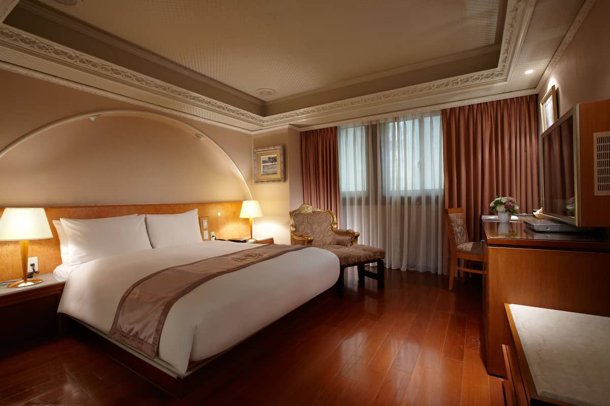 A hotel room with a large bed, two bedside lamps, a desk with a chair, and a large window with curtains.