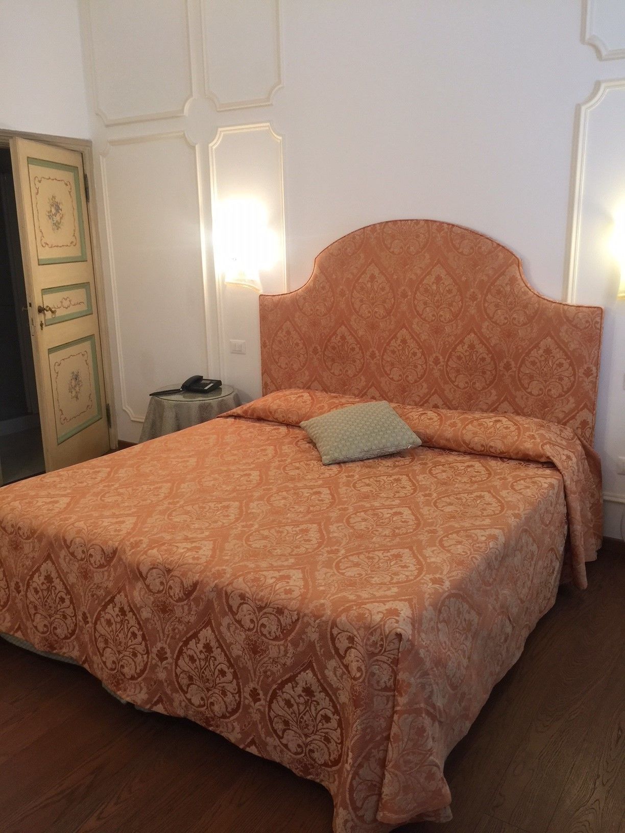 Una cama con cabecera naranja y ropa de cama estampada, una mesita de noche con lámpara, y piso de madera.