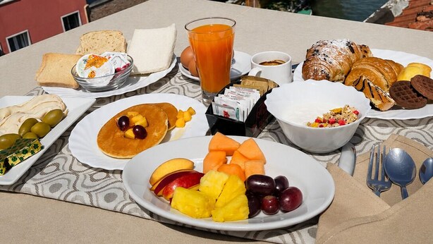 A breakfast table with various foods and a glass of orange juice, overlooking a canal with buildings and boats. - Ca' Dei Polo (Venice)