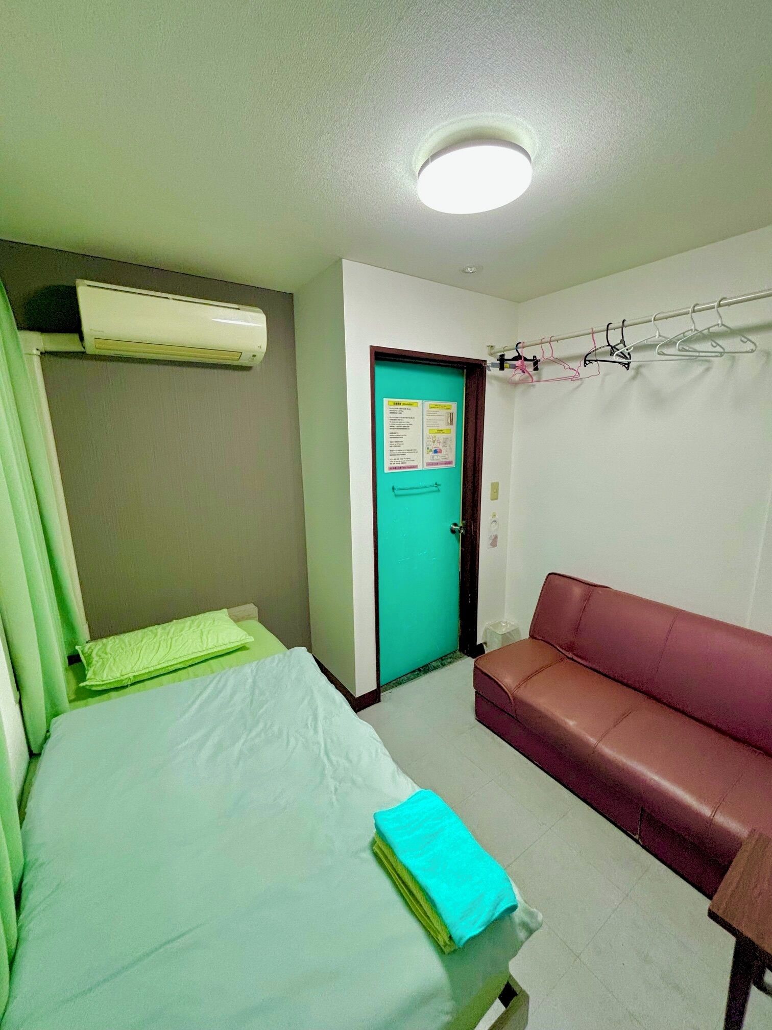 A room with a bed, a couch, an air conditioning unit, a door with a notice, and a coat rack.