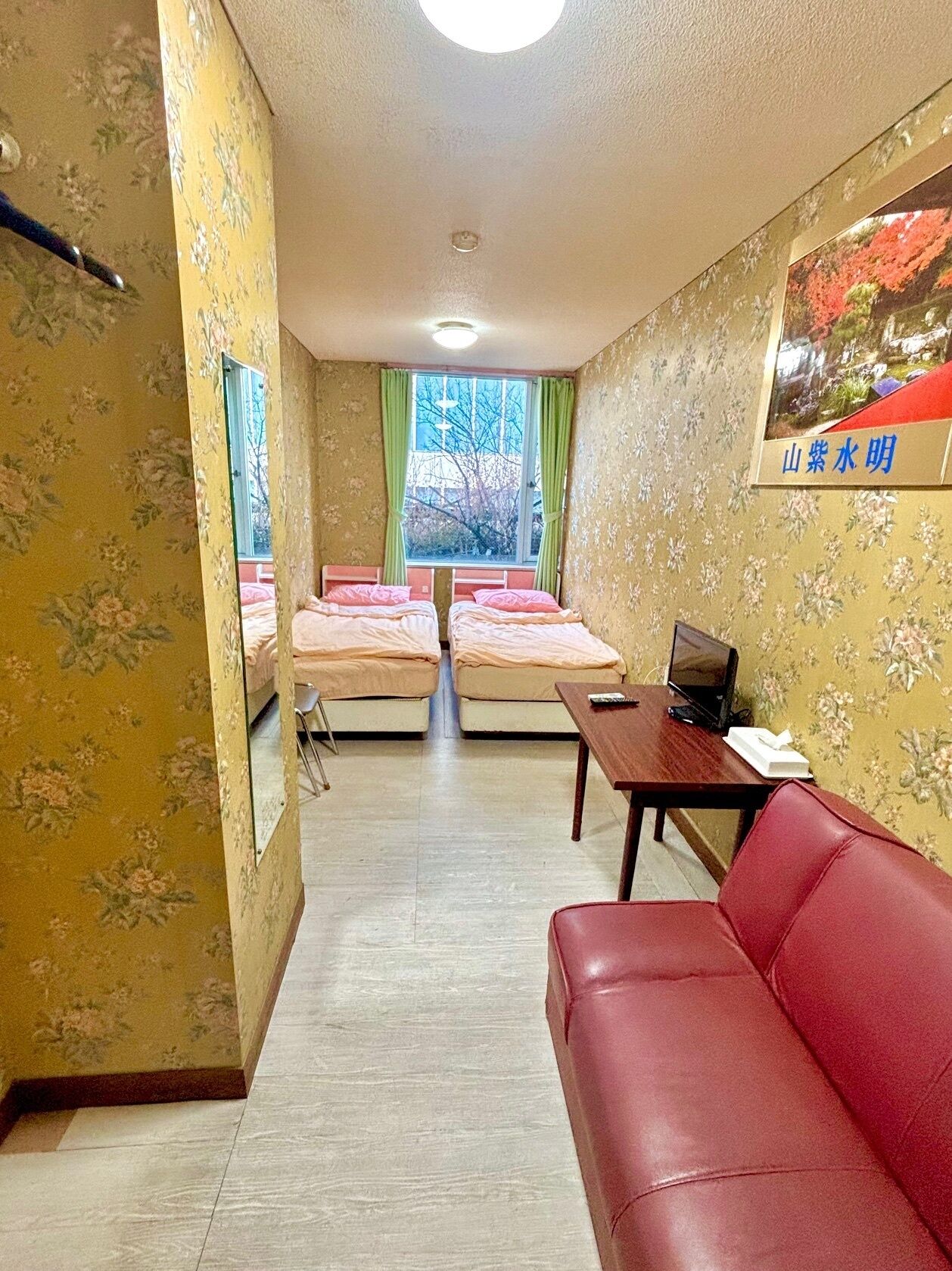 A hotel room with two beds, a red sofa, a desk with a laptop, and a window with green curtains.