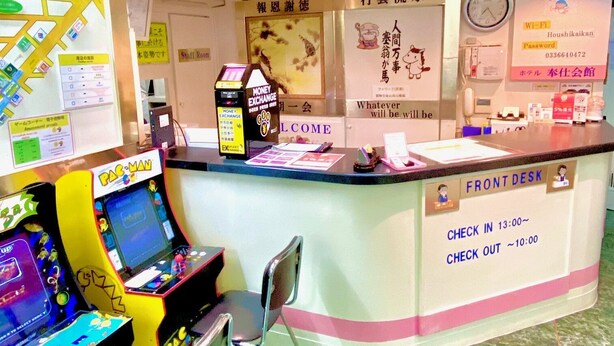 A counter area with a 'Money Exchange' machine, a 'Welcome' sign, and a 'Front Desk' sign indicating check-in and check-out times. - Hotel Hoshi Kai Kan (Tokyo)