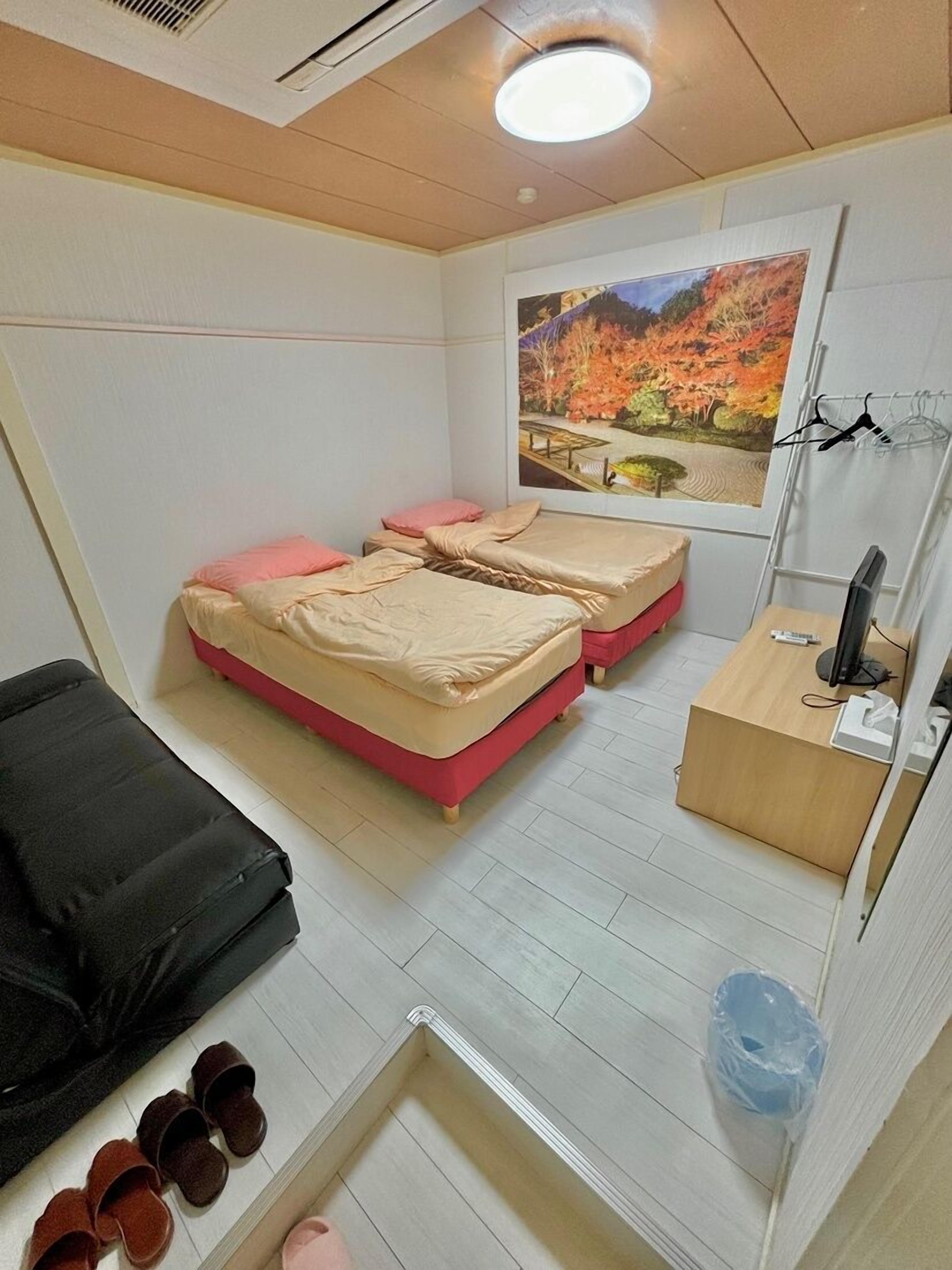 A small, compact room with two beds, a desk with a computer, and a window with a view of autumn trees.