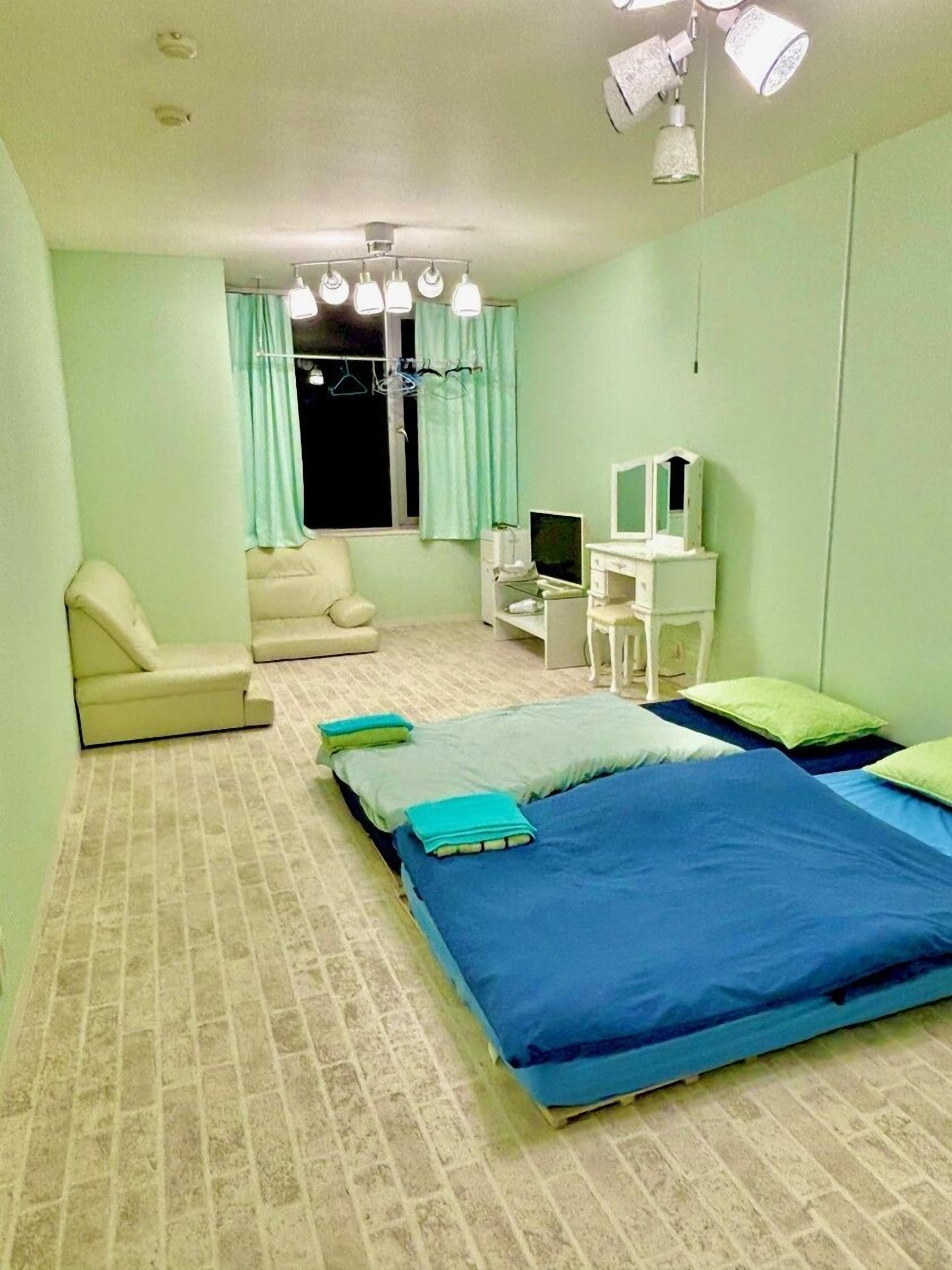 A bedroom with two beds, a chair, a desk with a computer, and a window with curtains.