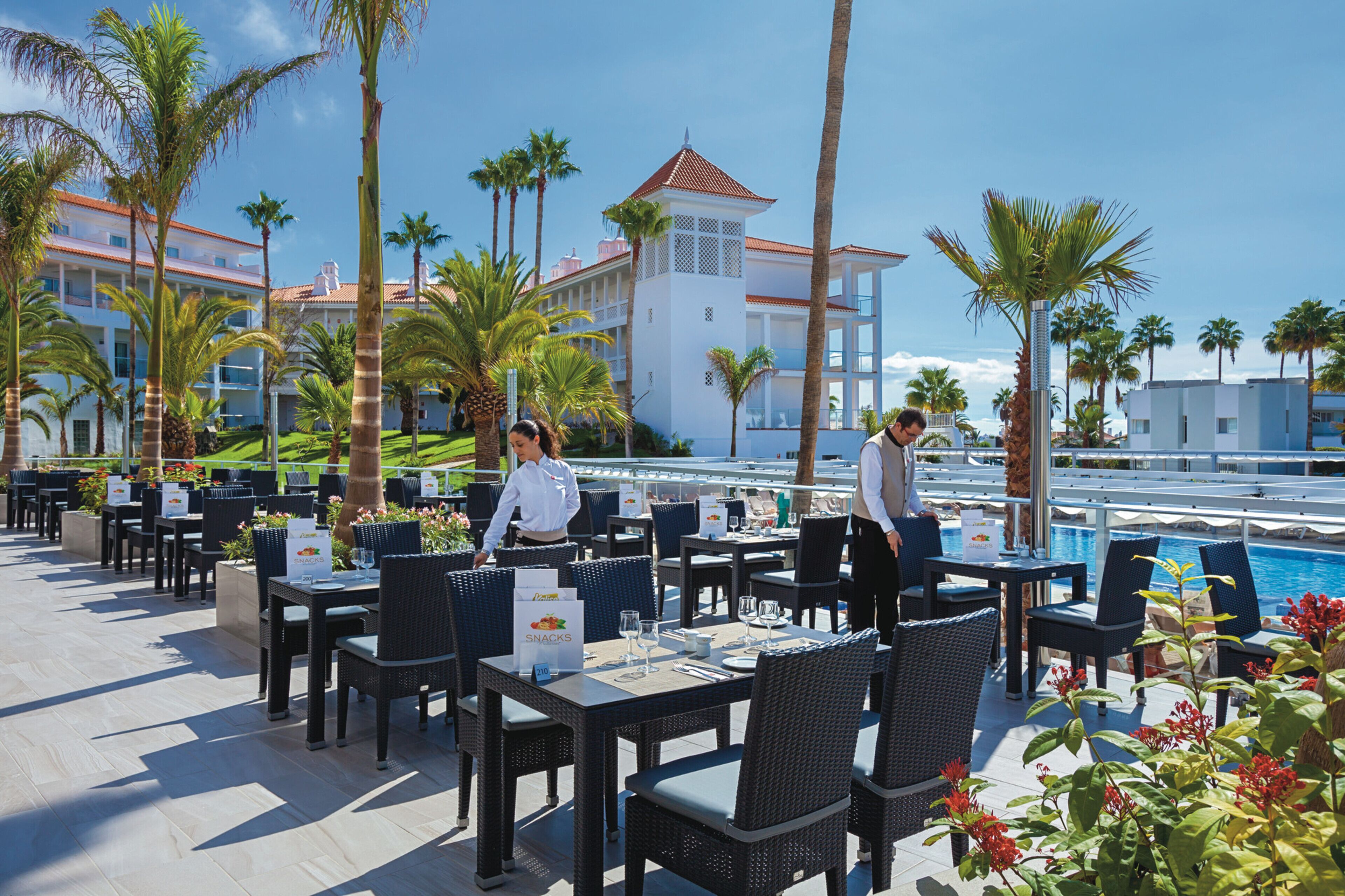 An outdoor dining area with tables and chairs, a pool, and palm trees.