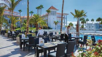 An outdoor dining area with tables and chairs, a pool, and palm trees.