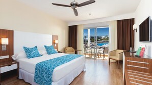 A hotel room with a large bed, a desk, a chair, a TV, and a balcony with a pool view. - Hotel Riu Arecas - Adults Only (Adeje)