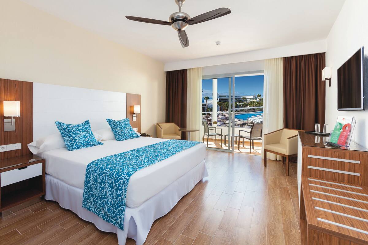 Camera d'hotel con un letto grande, una scrivania, una sedia, una TV e un balcone vista piscina.
