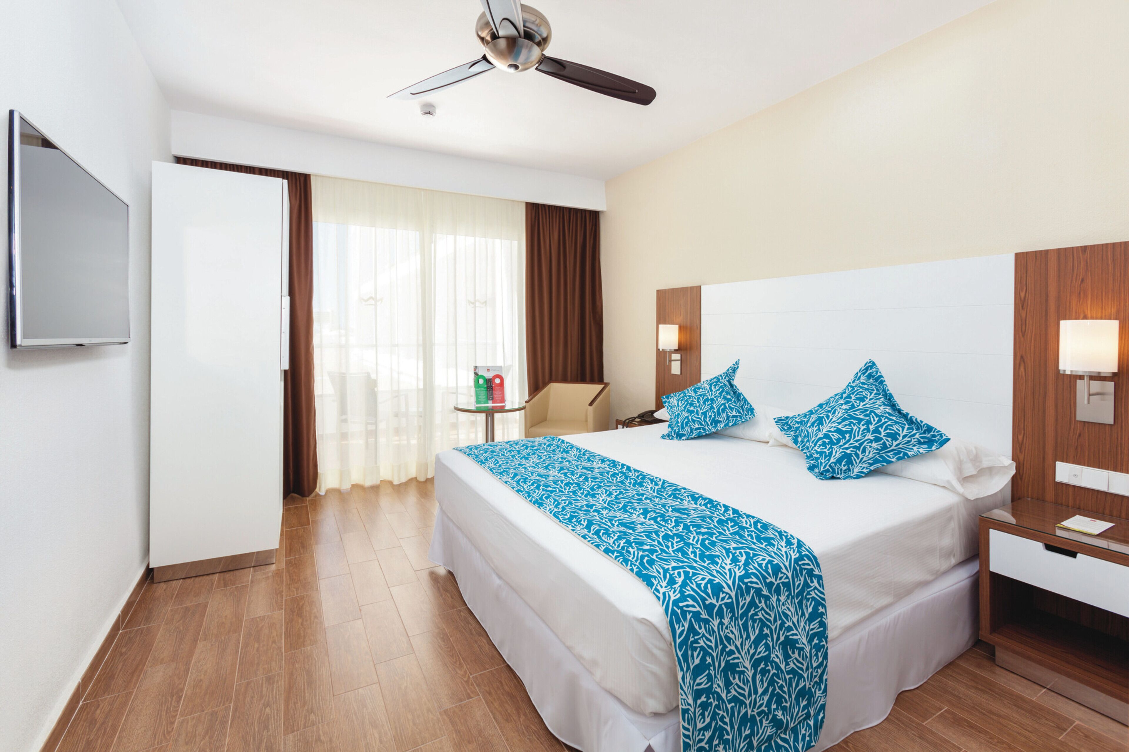 Una habitación de hotel con cama, ventilador de techo, televisor, mesita de noche y velador.