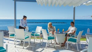 A covered outdoor seating area with white wicker furniture and a view of the ocean. - Hotel Riu Arecas - Adults Only (Adeje)